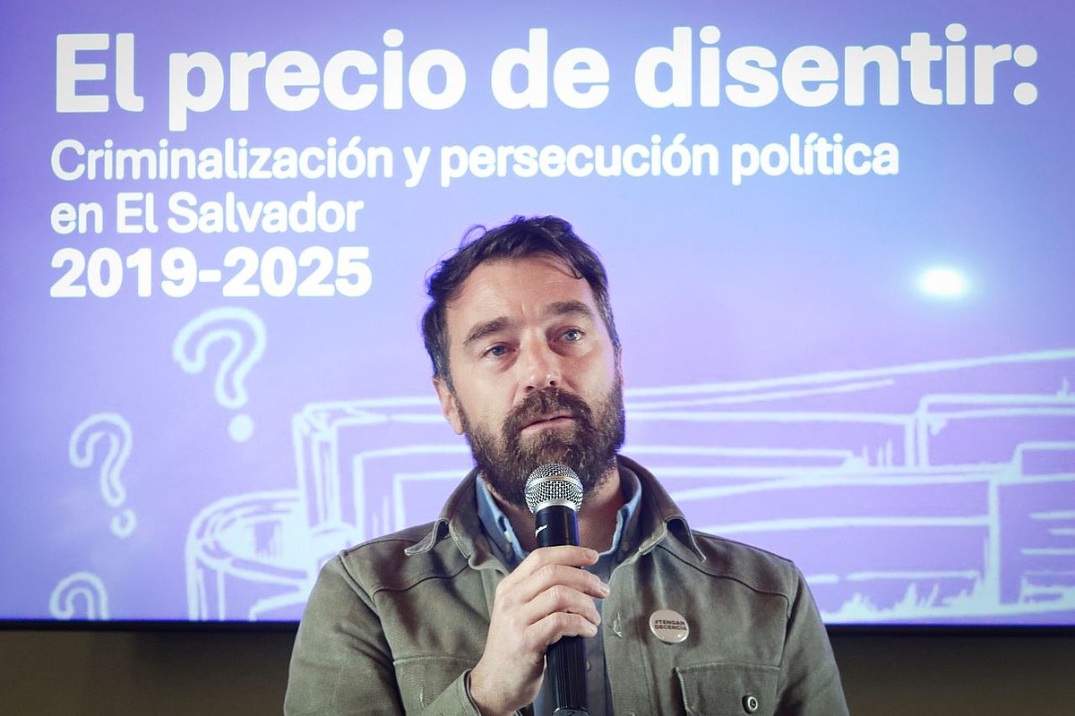 EIn Mann spricht in ein Mikrofon, hinter ihm eine Titelfolie einer Präsentation mit der Aufschrift "El precio de disentir - Criminalización y persecución política en El Salvador 2029-2025"