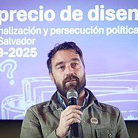 EIn Mann spricht in ein Mikrofon, hinter ihm eine Titelfolie einer Präsentation mit der Aufschrift "El precio de disentir - Criminalización y persecución política en El Salvador 2029-2025"