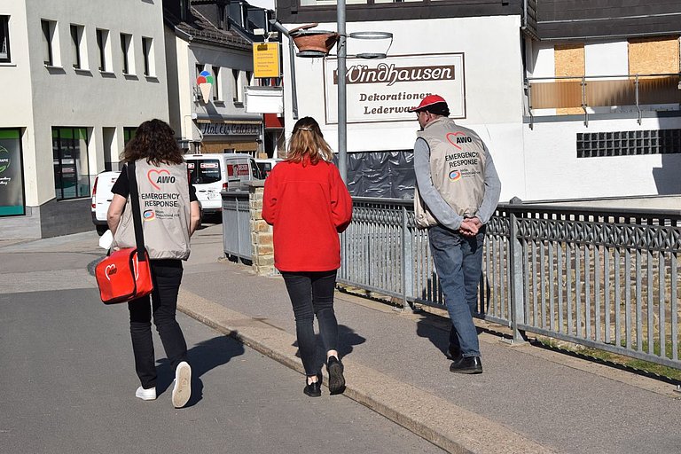 Zwei Frauen und ein Mann sind von hinten zu sehen. Sie laufen eine Straße entlang. Der Mann und die eine Frau tragen Westen mit der aufschrift "AWO - Emergency Respond".