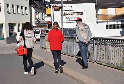 Zwei Frauen und ein Mann sind von hinten zu sehen. Sie laufen eine Straße entlang. Der Mann und die eine Frau tragen Westen mit der aufschrift "AWO - Emergency Respond".