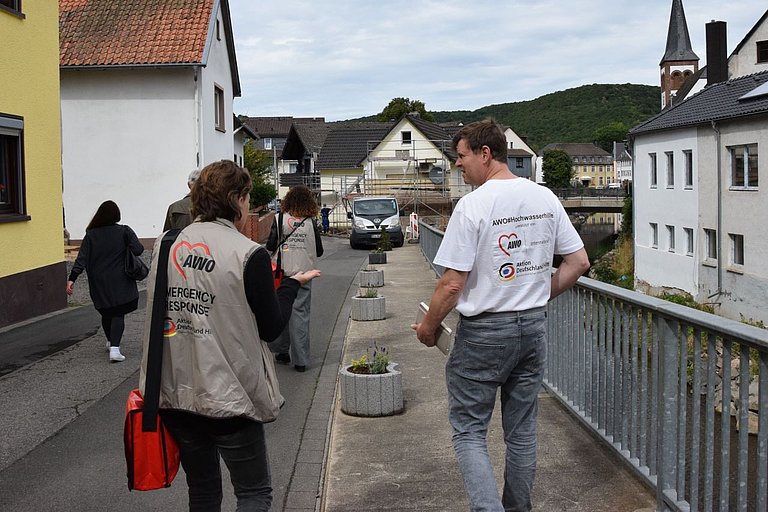 Ein Mann und eine Frau laufen durch eine Straße und unterhalten sich. Beide haben Logos auf der Kleidung. Dort steht "AWO International" und "Aktion Deutschland Hilft".