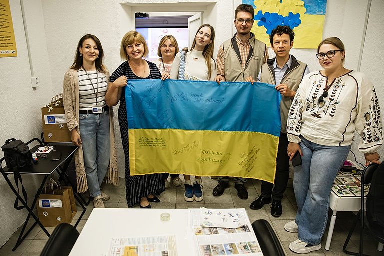 7 Frauen und Männer halten eine große ukrainische Flagge und schauen in die Kamera.