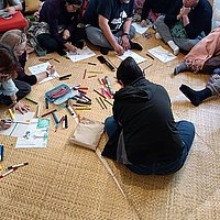 Eine Gruppe Personen sitzt auf dem Boden und malt, Stifte und Blätter liegen verteilt.