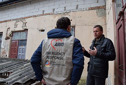 Zwei Männer stehen vor einem Haus und unterhalten sich. Auf der Weste des einen Mannes steht "AWO Emergency Response, Aktion Deutschland Hilft"