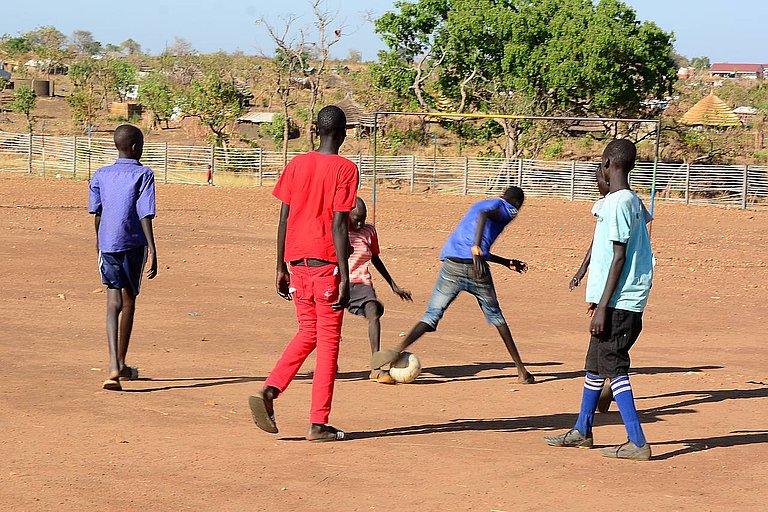 Titelbild der aktuellen weitblick-Ausgabe: Auf dem Weg zu unserem Hilfsprojekt im Adjumani Distrikt in Uganda spielen Jugendliche Fußball auf einem Feld. Das „Recht auf Spiel“ und ein faires Miteinander spielen auch in unserem Projekt eine große Rol