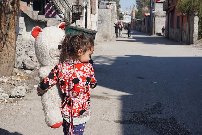 Ein Mädchen hält einen großen Teddybären in dem Arm und läuft eine Straße entlang
