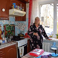 Eine ältere Frau steht in der Küche vor ihrem Fenster und lächelt in die Kamera.