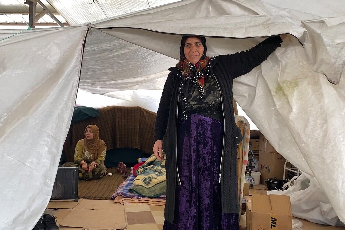A woman stands in the door of a tent. In the tent another woman sits on the floor.