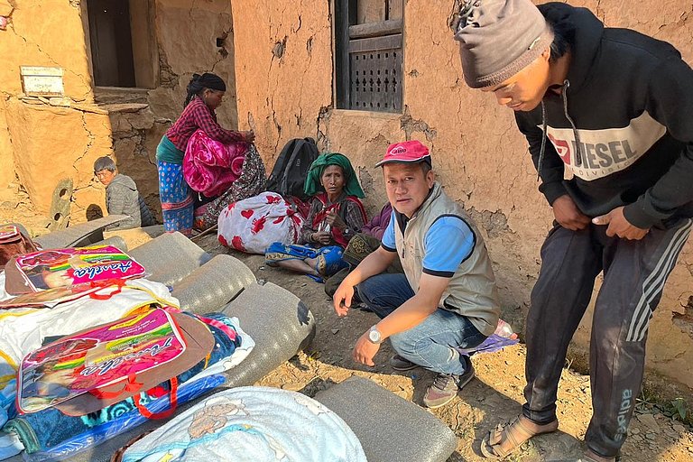 A man is sitting on the ground, with supplies next to him, and other people are handing out the supplies.