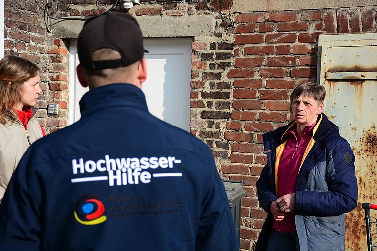 Ein Mann trägt eine Jacke mit der Aufschrift "Hochwasser-Hilfe". Er unterhält sich mit einer Frau, die vom Hochwasser 2021 betroffen war.