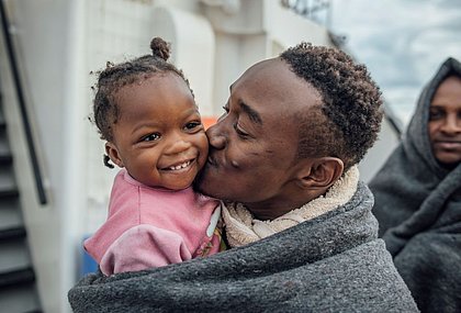 Ein Mann hält seine kleine Tochter im Arm. Er lächelt und gibt ihr einen Kuss auf die Wange.Beide stehen auf einem Schiff und sind in eine warme Decke eingewickelt.