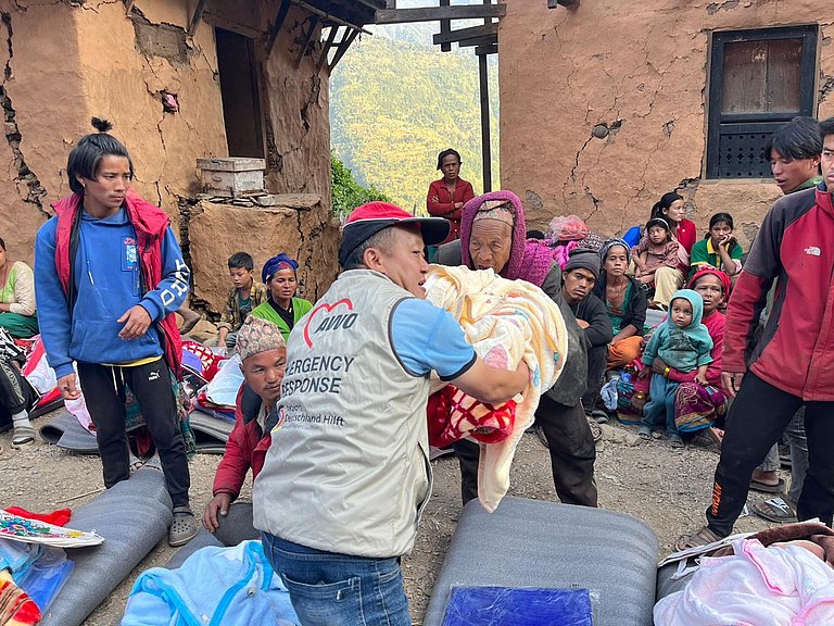 Man handing relief items to woman, with many children and young people in the background.