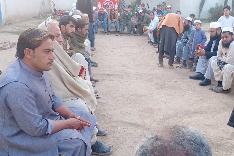 Refugees are sitting on benches in a circle outside. The gaze is directed downwards. Two people are walking around with blank forms in their hands. In the background, there are white walls from houses.