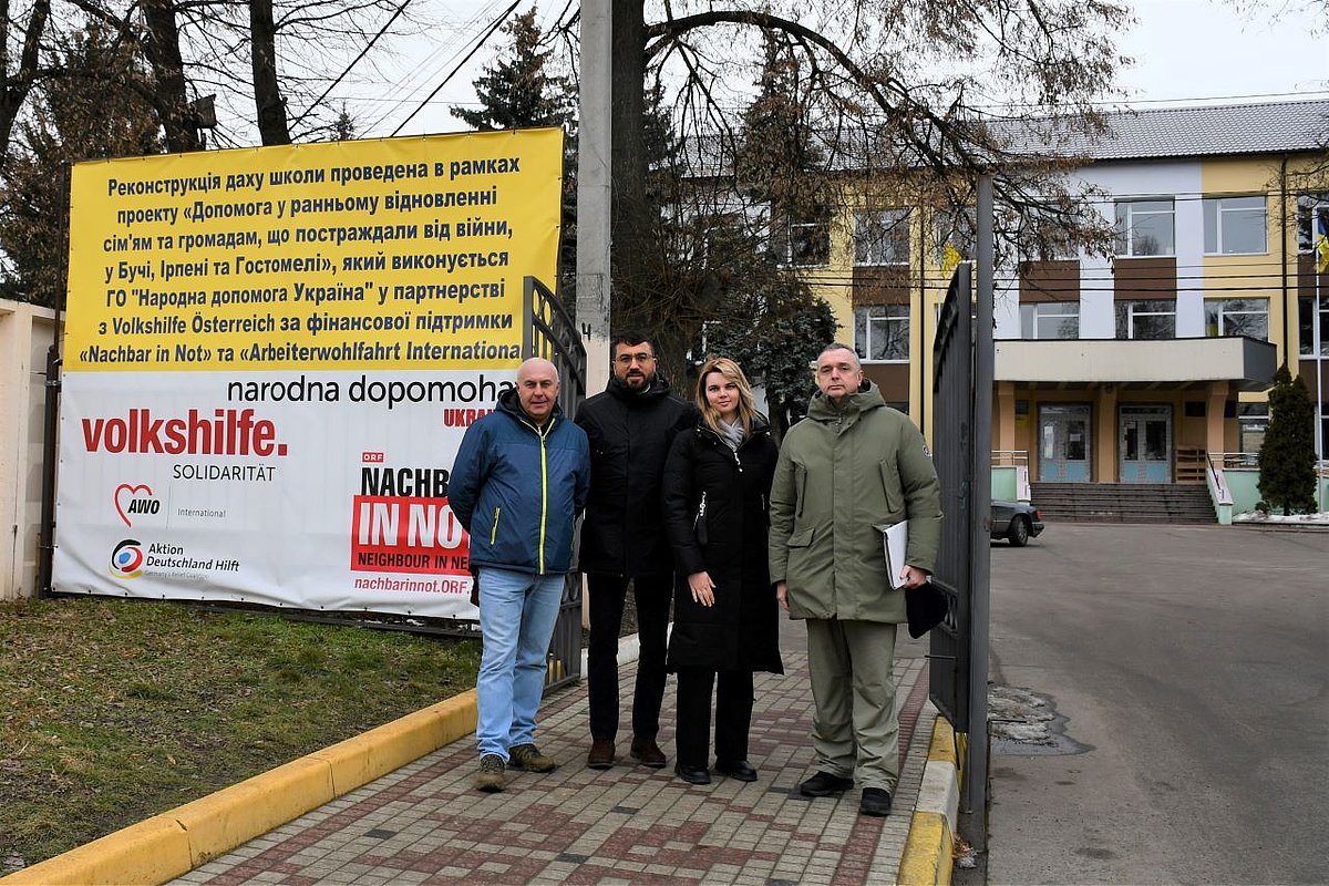 drei Männer und eine Frau stehen vor dem Tor einer Schule. Hinter ihnen ist ein großer Banner zu sehen mit den Logos der Volkshilfe, AWO International und Aktion Deutschland Hilft.