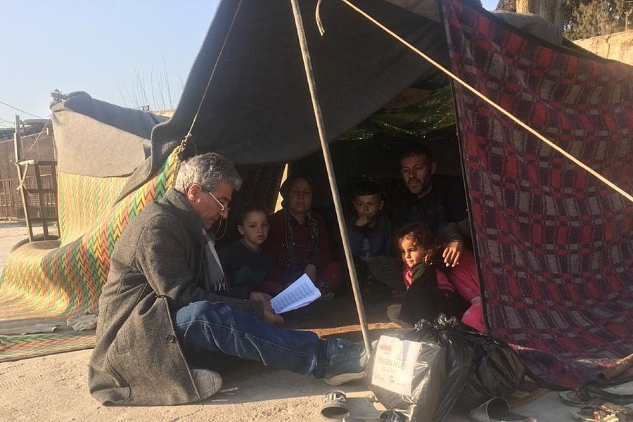Ein Mann sitzt vor einem Feld und hat eine Liste in der Hand. In dem Zelt sitzt eine Familie. Daneben liegt ein Plastikbeutel mit Hilfsgütern.