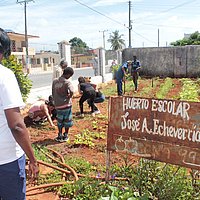 Auf einem Schild in einem Garten steht Schule José A Echeverria. Es steht eine Mitarbeiterin im Vordergrund, dahinter knien Kinder im Garten.