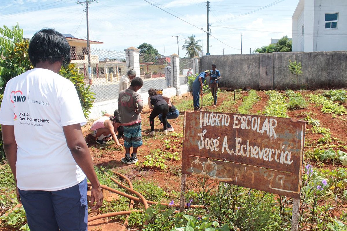 Auf einem Schild in einem Garten steht Schule José A Echeverria. Es steht eine Mitarbeiterin im Vordergrund, dahinter knien Kinder im Garten.