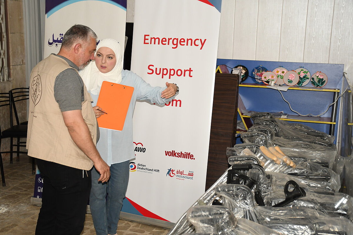 Eine Frau und ein Mann unterhalten sich. Im Hintergrund steht ein Banner mit der Aufschrift "Emergency Response". Neben den beiden stehen verpackte Rollstühle.