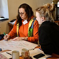 Two women. One is writing on a large sheet of paper.