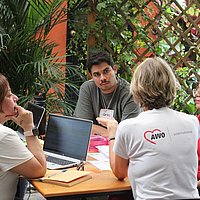 Vier Personen sitzen um einen Tisch mit Laptop und Arbeitsmaterialien herum und unterhalten sich.