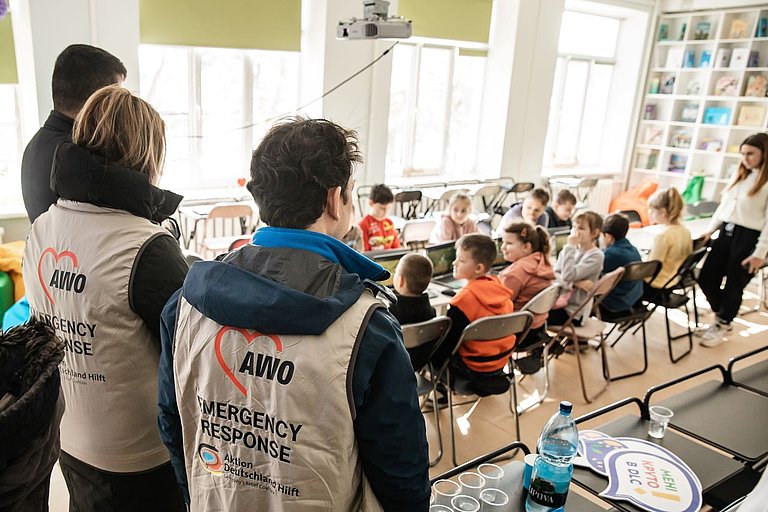 Ein Mann und eine Frau stehen in einem Klassenraum vor einer Gruppe Kinder, die an einem Tisch sitzt. Jedes Kind hat einen Laptop vor sich. Der Mann und die Frau tragen Westen mit dem Aufdruck: "AWO Emergency Response, Aktion Deutschland Hilft".