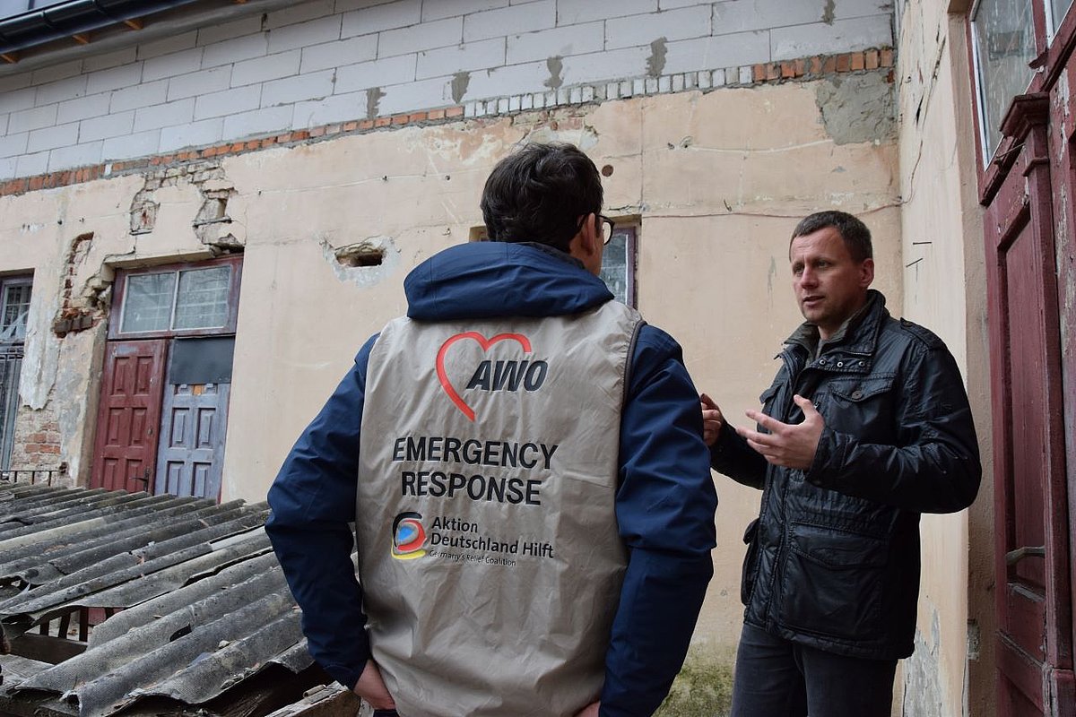 Zwei Männer stehen vor einem beschädigten Haus und unterhalten sich. Der eine Mann trägt eine Weste mit der Aufschrift "AWO, Aktion Deutschland Hilft, Emergency Response".