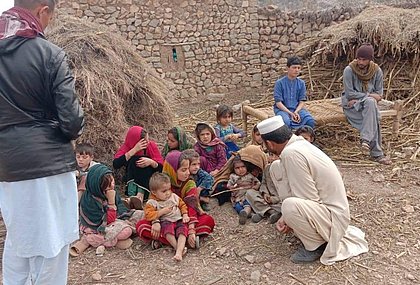 Children are sitting on the ground outside. Three men are also standing there talking to them. Straw and a stone wall can be seen in the background. 